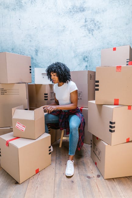 A woman with curly dark hair, dressed in a white t-shirt, blue jeans, and white sneakers, is sitting on a wooden chair surrounded by numerous cardboard boxes during a home relocation or packing process. She appears to be sealing or preparing a box, with some boxes closed and others open, revealing packing materials inside. The boxes are stacked and arranged on a light wooden floor within a room that has a textured light blue wall. Several boxes are secured with red and black packing tape, indicating organized packing for moving purposes. The scene captures the interior of a property, possibly during the final stages of a furniture transport and loading process, with the woman actively involved in packing or organising items for a professional removals service, such as those provided by Croydon Removals, for a house move in the Croydon area.
