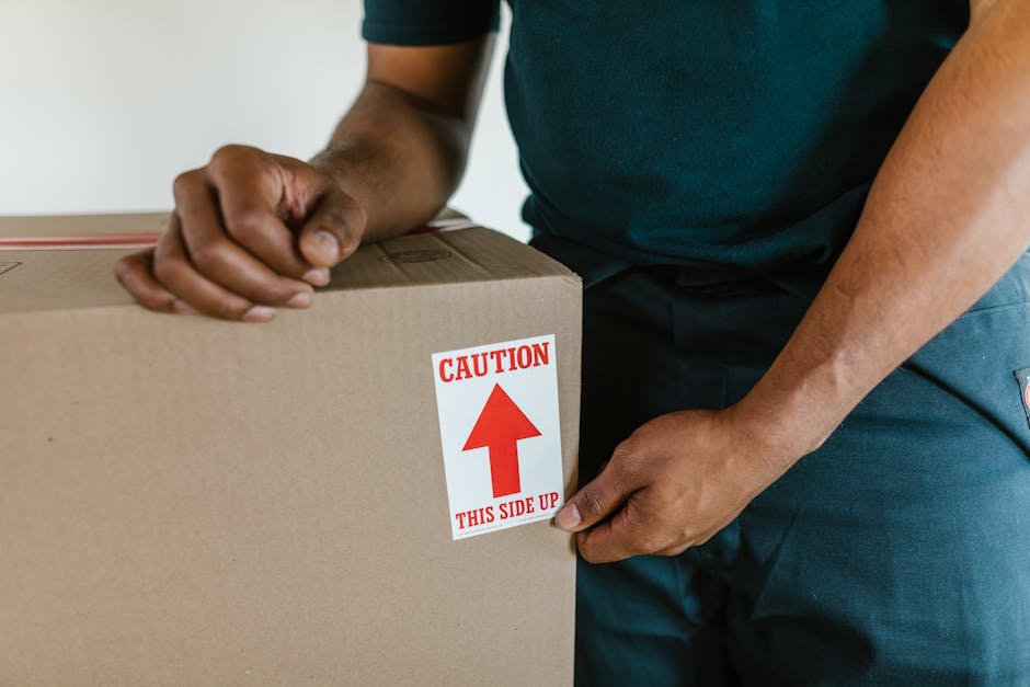 Close-up of a person wearing a dark blue shirt and matching trousers, positioning a cardboard moving box inside a property. The individual is gripping the box with both hands, one on the side and the other on the top corner, ensuring careful handling. The cardboard box is sealed with packing tape and features a bright red and white caution label that indicates 'Caution: This Side Up' with an upward-pointing arrow, highlighting safe orientation during the home relocation process. The background shows part of an indoor space with plain white walls, and the person appears to be preparing the box for transportation or loading onto a van. This image relates to furniture transport and packing procedures involved in professional removals services, such as those provided by Croydon Removals, ensuring secure handling of belongings during house moves, especially when navigating tight access areas during relocation in East Croydon.