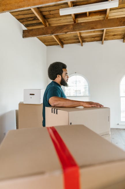 A woman with curly dark hair, dressed in a white t-shirt, blue jeans, and white sneakers, is sitting on a wooden chair surrounded by numerous cardboard boxes during a home relocation or packing process. She appears to be sealing or preparing a box, with some boxes closed and others open, revealing packing materials inside. The boxes are stacked and arranged on a light wooden floor within a room that has a textured light blue wall. Several boxes are secured with red and black packing tape, indicating organized packing for moving purposes. The scene captures the interior of a property, possibly during the final stages of a furniture transport and loading process, with the woman actively involved in packing or organising items for a professional removals service, such as those provided by Croydon Removals, for a house move in the Croydon area.