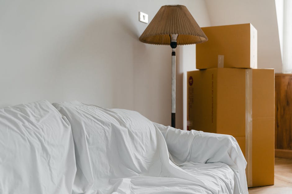 Inside a room prepared for house removals, several cardboard moving boxes are stacked against a wooden wall, with one box secured using clear packing tape. To the left, a white sofa covered with a protective sheet is positioned near a beige wall, and a tall floor lamp with a brown lampshade stands beside the boxes. The lighting appears natural, coming from a window outside the frame. The scene suggests the early stages of a home relocation process, with packing materials ready for transport and furniture prepared for the move. Croydon Removals, a professional moving company, can facilitate such furniture transport and packing services, ensuring smooth house relocations. The space is inside a residential property, with visible details of packing and loading preparations in progress.
