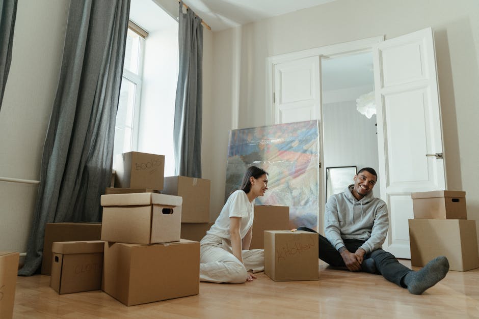 A young woman and a man are sitting on a light wooden floor inside a room with cream walls and a high ceiling, surrounded by packed cardboard moving boxes, some labeled with handwritten words such as 'books' and 'clothes'. The woman is wearing a white T-shirt and beige pants, and is sitting cross-legged on the floor, looking at the man with a smile. The man, dressed in a grey hoodie and dark joggers, is also seated on the floor, smiling at the woman. Behind them, there are large windows with grey curtains letting in natural light, and a white open door revealing another part of the house. A large, partially visible canvas painting leans against the wall near the window, along with a framed mirror or picture. The scene captures the initial stages of a home relocation process, possibly involving furniture transport and packing, with the presence of several packed boxes and packaging materials indicating preparation for moving day, consistent with house removals services offered by Croydon Removals.