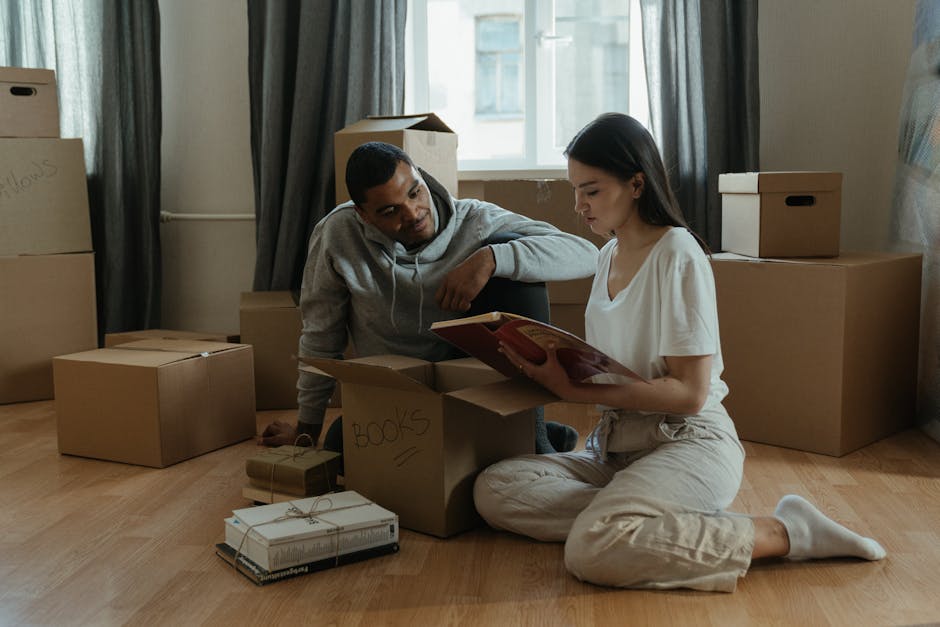 Inside a room prepared for house removals, several cardboard moving boxes are stacked against a wooden wall, with one box secured using clear packing tape. To the left, a white sofa covered with a protective sheet is positioned near a beige wall, and a tall floor lamp with a brown lampshade stands beside the boxes. The lighting appears natural, coming from a window outside the frame. The scene suggests the early stages of a home relocation process, with packing materials ready for transport and furniture prepared for the move. Croydon Removals, a professional moving company, can facilitate such furniture transport and packing services, ensuring smooth house relocations. The space is inside a residential property, with visible details of packing and loading preparations in progress.